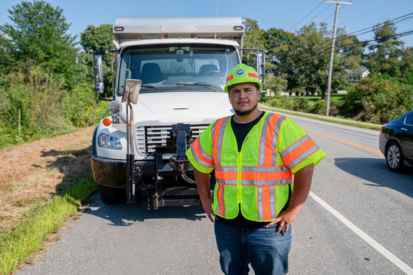 Roadside maintenance worker wearing a high‑visibility safety vest and a hard hat stands on the side of a road in front of a large white work truck. The truck is parked partially on the road near a grassy shoulder. The truck has tools mounted on its front, another vehicle is visible driving past on the right side of the road. Trees, power lines, and a sunny sky form the background.